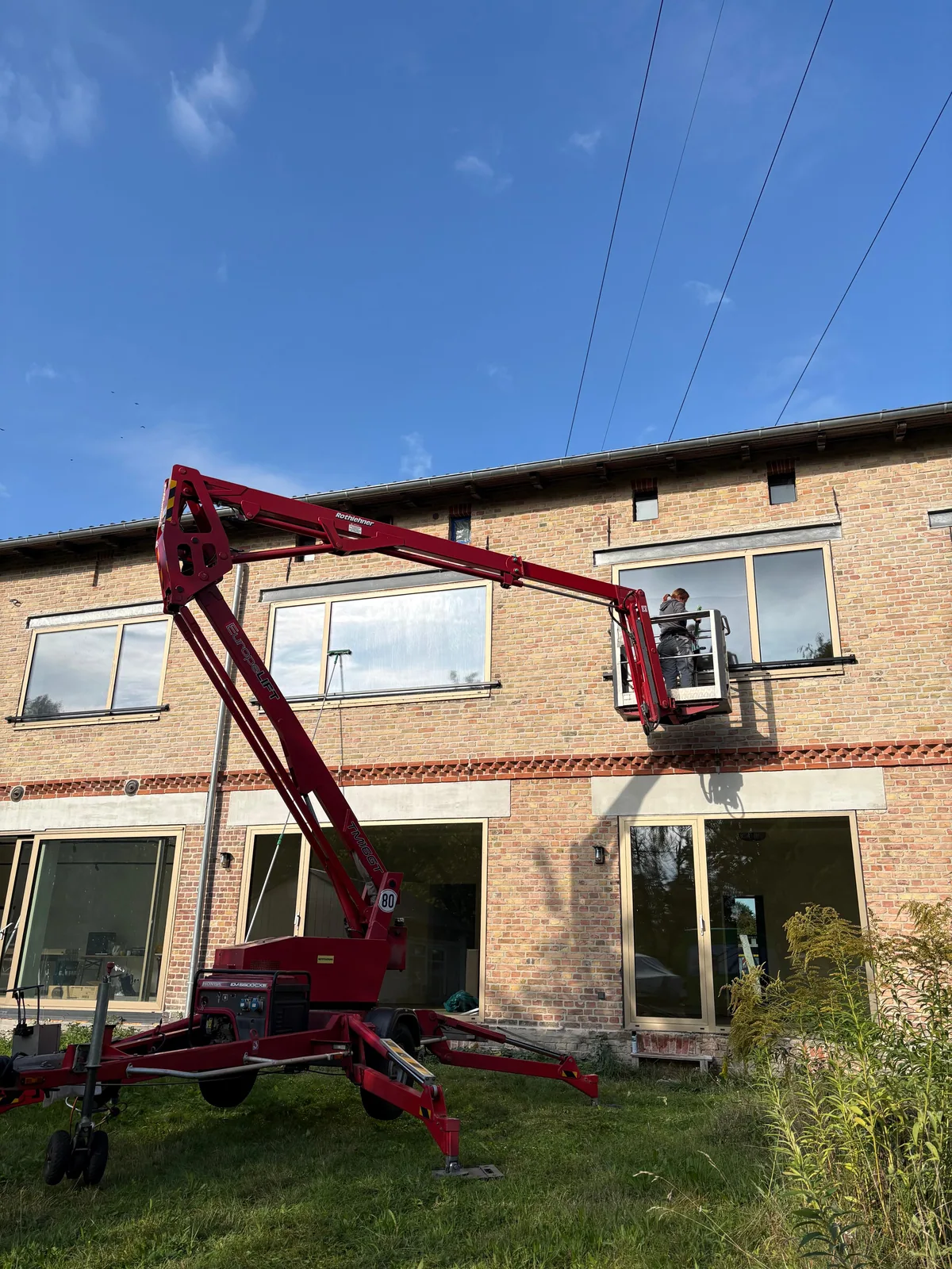 Ausgefahrene rote Arbeitsbühne an einer Klinkerfassade, ein Mitarbeiter der Wilke Reinigung reinigt vom Korb aus ein Fenster im Obergeschoss